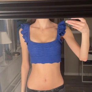 Blå croppad stickad topp med volang - Säljer en blå croppad stickad topp med breda axelband och snygga volangdetaljer. Toppen har en fyrkantig urringning och ett ribbat mönster över hela. Perfekt för dig som vill ha en unik och trendig look i sommar.