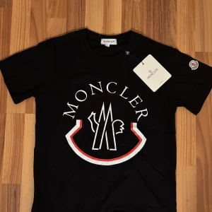 Moncler t-shirt - Svart t-shirt från Moncler med stor vit och röd logga framtill. Klassisk rund halsringning och korta ärmar. Ikonisk Moncler-patch på ärmen och mjukt bomullsmaterial. Perfekt för dig som gillar streetwear och vill sticka ut med en snygg logga.