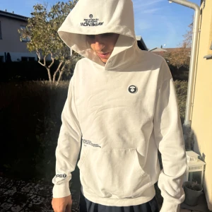 Vit hoodie från AAPE med tryck - Säljer en clean vit hoodie från AAPE med svart logotyptryck på bröstet, huvan, axeln och ärmen. Hoodien har stor huva, känguruficka och ribbade muddar. Perfekt för dig som gillar streetwear och vill ha något som sticker ut.