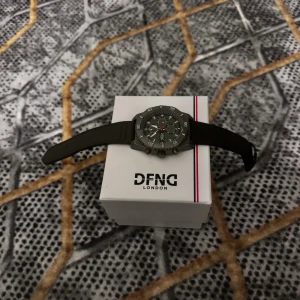 Svart sportig klocka från DFNG London - Snygg och sportig klocka från DFNG London med svart armband och rund urtavla. Klockan har flera små urtavlor, tydliga siffror och detaljer i silver. Perfekt accessoar för dig som gillar en stilren och modern look.