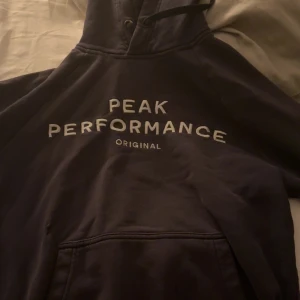 Mörkblå hoodie från Peak Performance - Säljer en mörkblå hoodie från Peak Performance med vit logga framtill. Klassisk modell med huva, känguruficka och ribbade muddar. Perfekt för chill dagar eller när du vill ha en sportig look.