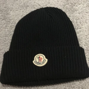 Svart ribbad Moncler mössa - Svart ribbstickad mössa från Moncler med klassisk uppvikt kant och ikonisk Moncler-logga framtill. Mössan är tillverkad i mjukt material som håller dig varm och har en stilren look som funkar till alla jackor. Perfekt för dig som vill ha en snygg och enkel mössa med premiumkänsla.