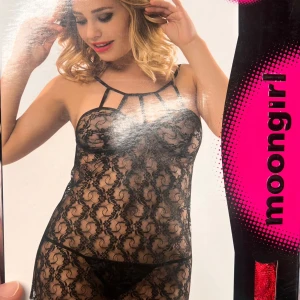 Svart spetsklänning från Moongirl - Svart, transparent kortklänning i spets med tunna axelband och dekorativa banddetaljer framtill. Klänningen har ett blommigt spetsmönster och är figurnära med en sexig look. Perfekt för dig som vill ha något extra till speciella tillfällen.