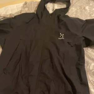 Svart vindjacka från Haglöfs med huva - Svart vindjacka från Haglöfs med huva och hel dragkedja framtill. Jackan har två snedställda fickor med dragkedja och en diskret logga på bröstet. Perfekt för blåsiga dagar och outdoor-äventyr. Lätt och smidig design.