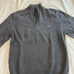 Grå stickad tröja från Gant - Snygg grå stickad tröja från Gant med halv dragkedja och ribbad krage. Klassisk modell med broderad Gant-logga på bröstet. Perfekt för lager-på-lager och passar till både jeans och chinos.