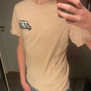 Beige Nike t-shirt med tryck - Säljer en beige t-shirt från Nike med svartvitt tryck på bröstet och stort tryck på ryggen. Klassisk passform med rund hals och korta ärmar. Skön och lätt i bomull, perfekt till jeans eller shorts.