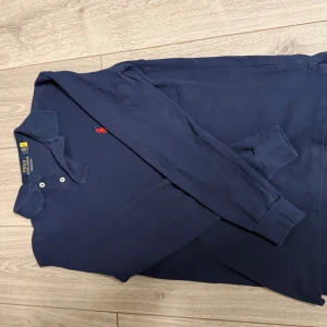 Marinblå långärmad pikétröja Polo Ralph Lauren - Snygg marinblå långärmad pikétröja från Polo Ralph Lauren med röd broderad logga på bröstet. Tröjan har klassisk krage, knappar vid halsen och är i bomull med slim fit-passform. Perfekt för en stilren och sportig look.