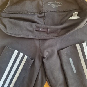 Svarta Adidas leggings med vita ränder - Svarta leggings från Adidas med klassiska vita ränder längs sidorna och Adidas-logga på låret. Tillverkade i stretchigt Climalite-material som andas, perfekt för träning. Tight passform och hög midja för en sportig look.