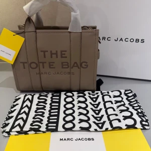 Beige tote bag från Marc Jacobs Medium - Snygg beige tote bag från Marc Jacobs i äkta skinn med präglad logga framtill. Väskan har robusta handtag, guldfärgade metalldetaljer och dragkedja upptill. Insidan är fodrad med mörkt tyg och har en innerficka. Perfekt storlek för dina essentials.