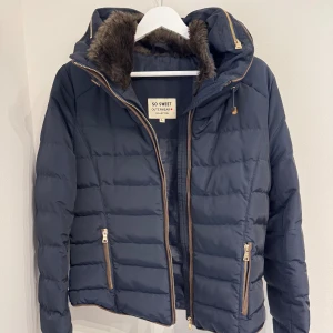 Mörkblå pufferjacka quiltad  - Very good condition except the zipper, it is not working. The size is L but it runs small and better for M. Mörkblå pufferjacka från So Sweet med fuskpäls i luvan och flera guldfärgade dragkedjor. Jackan har quiltad design, lång ärm och två fickor framtill. Perfekt för kalla dagar med sin varma och mysiga känsla.
