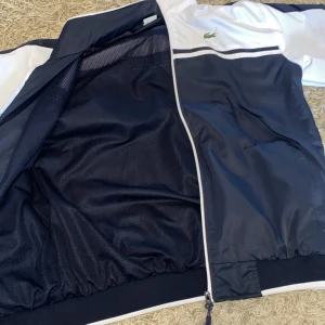 Blåvit vindjacka från Lacoste Sport - Snygg vindjacka från Lacoste Sport i marinblått och vitt med klassisk krokodillogga på bröstet och broderad Lacoste-text på ryggen. Jackan har hel dragkedja, ribbade muddar och sportig design med kontrastpaneler. 