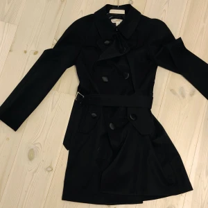 Svart trenchcoat H&M Comme des Garçons - Svart trenchcoat från H&M Comme des Garçons med klassisk krage, stora knappar och bälte i midjan. Långärmad modell i slät polyester med dubbla knapprader och fickor framtill. Perfekt för dig som gillar stilrena och tidlösa plagg.