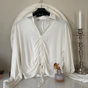 Bodyflirt topp - Supersnygg vit skjorta från Bodyflirt med lång ärm och krage. Framsidan har cool snörning och man kan dra åt snöre  och rynkade detaljer som ger en unik look. Skjortan är gjord i mjukt syntetmaterial och har en omlottstil med v-ringning. Perfekt för dig som gillar trendiga detaljer och stilren design.