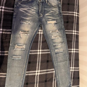 Blå slitna skinny jeans, storlek 32 - Säljer ett par blå skinny jeans med slitningar och trasiga detaljer framtill. Jeansen har klassisk femficksdesign, normal midja och är tillverkade i 85% bomull och 15% polyester. Perfekta för dig som gillar en edgy och trendig look.