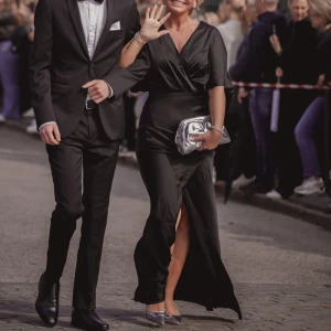 Svart lång omlottklänning med slits - Säljer en svart lång omlottklänning i glansigt material med v-ringning, vida ärmar och öppen rygg. Klänningen har en snygg slits framtill och markerad midja. Perfekt för bal. Stilren och elegant look.