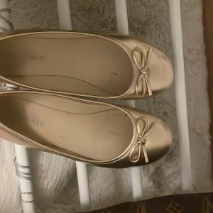 Guldiga ballerinaskor med rosett - Snygga ballerinaskor i metallic guld från Anna Field. Skorna har rund tå, platt sula och en söt rosettdetalj framtill. Perfekta för dig som vill addera lite extra bling till din outfit. Använt ungefär 2-3 gånger 