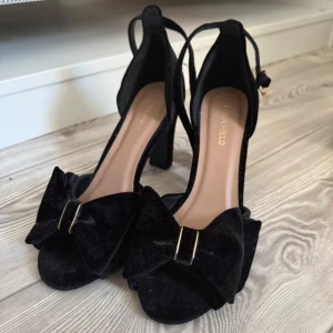 Svarta sammetsklackar med rosett - Snygga svarta klackar från Anna Field i mjuk sammet med bred klack och öppen tå. Skorna har en stor dekorativ rosett framtill och justerbar rem runt vristen med guldfärgat spänne. Perfekta för dig som vill sticka ut med en elegant och trendig look. Använda 1 gång