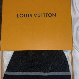 Lv mössa  - Snygg svart mössa från Louis Vuitton med diskret monogrammönster och grå kant. Tillverkad i mjuk ull som håller dig varm och stilren under kalla dagar. Perfekt accessoar för dig som gillar exklusiva märken och klassisk design.