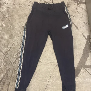 Mörkblå mjukisbyxor från Hugo Boss - Svarta mjukisbyxor från Hugo Boss med vit HUGO-logga på vänster lår och logoband längs båda sidorna. Byxorna har snörning i midjan och är tillverkade i mjukt bomullsmaterial. Perfekta för chill eller träning.