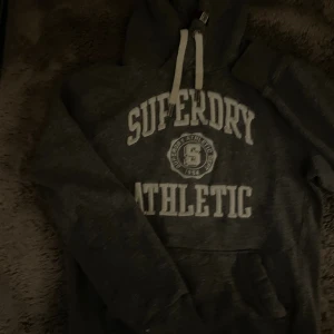 Mörkgrå Superdry hoodie med tryck - Mysig mörkgrå hoodie från Superdry med vit text 'Superdry Athletic' och emblem på bröstet. Tröjan har huva med vita snören och en stor magficka. Perfekt för chill dagar eller när du vill ha en sportig look.