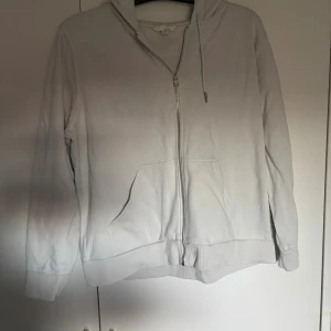 Vit hoodie med dragkedja från H&M - Säljer en stilren vit hoodie från H&M i storlek L. Tröjan har huva med snörning, dragkedja framtill och en stor känguruficka. Perfekt för chill dagar eller när du vill ha en enkel och clean look.