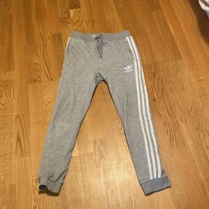 Grå mjukisbyxor från Adidas - Snygga grå mjukisbyxor från Adidas med klassiska vita ränder längs sidorna och logga på vänster ben. Byxorna har snörning i midjan, muddar vid bensluten och är gjorda i mjukt bomullsmaterial. Perfekta för chill eller träning.