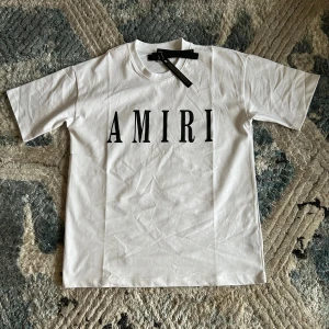 Vit AMIRI t-shirt med tryck - Snygg vit t-shirt från AMIRI med stort svart logotryck på bröstet. Klassisk rund hals och korta ärmar. Tillverkad i mjuk bomull som känns skön mot huden. Perfekt för dig som gillar stilrena och trendiga plagg med tydlig branding. Kom med bud! Endast använd en gång