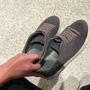 Grå sneakers från Axel Arigato - Snygga grå sneakers från Axel Arigato i mocka med vita sulor och matchande grå snörning. Skorna har diskret guldfärgad logga på sidan och detaljer i läder vid hälen. Perfekta för en clean och stilren look.