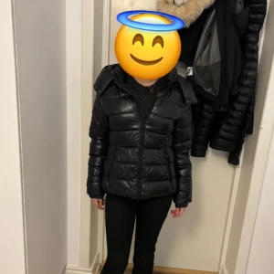 Svart pufferjacka med huva - Säljer en svart pufferjacka med huva och dragkedja framtill. Jackan har quiltad design och två fickor med dragkedja. Perfekt för kyliga dagar och enkel att matcha med olika outfits.