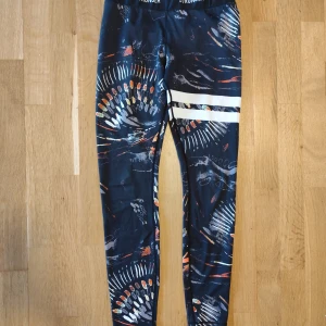 Mönstrade leggings från Stronger - Snygga leggings från Stronger med coolt abstrakt mönster i mörkblått, vitt, orange och gult. De har två vita ränder på ena benet och elastisk midja med logga. Perfekta för träning eller chill. Materialet är stretchigt och följsamt syntet.