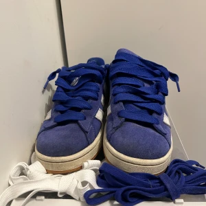Blå Adidas Campus sneakers mocka - Säljer ett par blå Adidas Campus sneakers i mocka med vita klassiska ränder och vit logga på plösen. Skorna har blå snören och medföljer även ett par vita snören. Snygg retrostil med låg profil och platt sula, perfekt för dig som gillar streetwear.