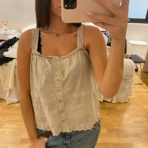 Beige blus med spets och knappar - Säljer en söt beige blus med breda axelband och fina spetsdetaljer. Blusen har knappar framtill och broderade kanter längst ner. Luftig och perfekt för varma dagar, passar till jeans eller kjol. Fräsch och feminin stil!