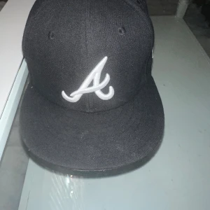 Svart Atlanta  keps - Säljer en svart keps från New Era med Atlanta Braves-logga broderad i vitt framtill. Baksidan har broderad text 'Greetings from Atlanta' och gröna vägskyltar. Klassisk rak skärm och snygg streetstil, perfekt för dig som gillar sport och urban look.