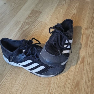 Svarta Adidas sneakers strl 36 - Säljer ett par svarta Adidas sneakers med klassiska vita ränder på sidorna. Skorna har snörning och är tillverkade i syntetmaterial och mesh för bra andningsförmåga. Perfekta för idrotten eller som bekväma skor till vardagen.