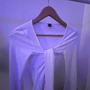 Vit långärmad topp med knyt - Säljer en vit långärmad topp från SHEIN med snygg knytning framtill. Toppen har en v-ringad hals och är gjord i mjukt, stretchigt material som sitter skönt mot kroppen. Perfekt för dig som gillar stilrena och trendiga plagg. så snygg jag nästan inte vill sälja den men den är för stor🤍🤍