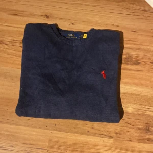 Marinblå stickad tröja Polo Ralph Lauren - Säljer en marinblå stickad tröja från Polo Ralph Lauren med klassisk röd logga broderad på bröstet. Tröjan har rund halsringning, ribbade muddar och är långärmad. Perfekt för dig som gillar stilren och tidlös design.