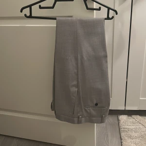 Grå kostymbyxor med pressveck - Snygga grå kostymbyxor med klassiskt pressveck och uppvikta benslut. Byxorna har diskreta fickor bak med knapp och är perfekta för en clean och stilren look. Materialet känns mjukt och har en lätt glansig finish. Storlek 29-32