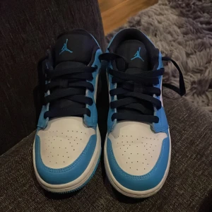 Nike Air Jordan 1 Low UNC blå/vit - Säljer ett par Nike Air Jordan 1 Low i den ikoniska UNC-färgen. Skorna har en snygg kombination av blått och vitt skinn, svarta swooshes och mörkblå snörning. Klassisk Jordan-logga på hälen och Jumpman på plösen. Perfekta sneakers för dig som vill sticka ut.