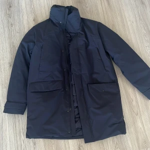 Mörkblå lång dunjacka från Dressmann - Säljer en stilren mörkblå dunjacka från Dressmann. Jackan är lång med hög krage, stora fickor framtill och stängs med både dragkedja och knappar. Yttermaterialet är slätt och vattenavvisande, perfekt för kalla dagar. Insidan är quiltad för extra värme. Använd 1 gång! Nypris runt (1000kr).