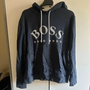 Hugo boss hoodie  - Snygg svart hoodie från Hugo Boss med stort BOSS-tryck i vitt på bröstet. Tröjan har huva med vita snören och en klassisk känguruficka framtill. Perfekt för dig som gillar stilrena och bekväma plagg med tydlig logga.