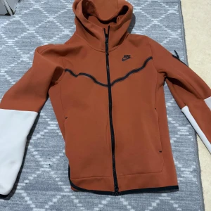 Nike brun hoodiejacka XS - Nike hoodiejacka i brun färg med svarta detaljer och vita partier på ärmarna. Jackan har hel dragkedja, huva och två fickor framtill. Perfekt för dig som gillar sportig och clean stil. Köpte den för 1700kr och säljer den för 800