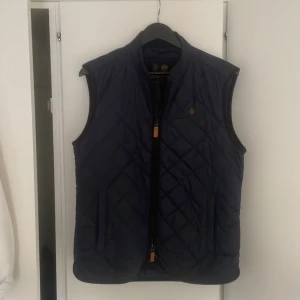 Mörkblå quiltad dunväst från Morris - Quiltad mörkblå dunväst från Morris med dragkedja framtill och diskret logga på bröstet. Västen har klassisk passform och snyggt rutmönster över hela ytan. Perfekt för lager-på-lager och stilren look.