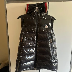 Moncler väst  - Snygg svart dunväst från Moncler med glansig finish och klassisk logga på bröstet. Västen har två dragkedjefickor, hög krage med röd och vit detalj samt quiltad design. Perfekt för lager-på-lager och streetstyle.