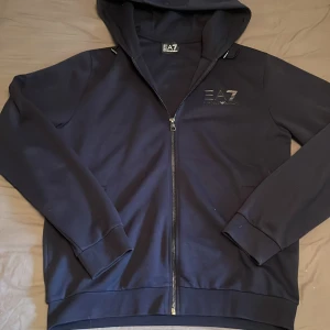 Mörkblå EA7 hoodie med dragkedja - Snygg mörkblå hoodie från EA7 Emporio Armani med huva och hel dragkedja framtill. Tröjan har diskret logga på bröstet och är tillverkad i mjukt material med långa ärmar och ribbade muddar. Perfekt för en chill och stilren look.