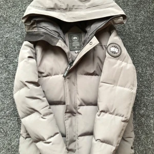 Canada Goose Macmillan jacka - Säljer en grå pufferjacka från Canada Goose med huva och klassisk logga på ärmen. Jackan har dragkedja framtill och är vadderad för extra värme. Perfekt för vintern. OBS! Priset kan diskuteras. Jackan har ett litet hål längst ner men syns ej och enkelt att fixa