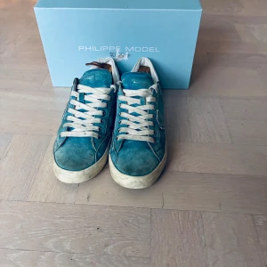 Philippe Model Junior - Snygga turkosa sneakers från Philippe Model med vit snörning och detaljer. Skorna har en rund tå, platt sula och är tillverkade i mocka med inslag av skinn. Vit häl med svarta prickar och klassisk logga på sidan. Perfekt för dig som gillar stilrena och unika sneakers. Priset är inte hugget i sten!