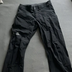 Haglöfs byxor - Svarta täckbyxor från Haglöfs i herrmodell, storlek S passa M också. Byxorna har flera praktiska fickor med dragkedja och bälteshällor. Materialet är slitstarkt och passar perfekt för utomhusaktiviteter. Snygg minimalistisk design med diskret Haglöfs-logga på benet. 