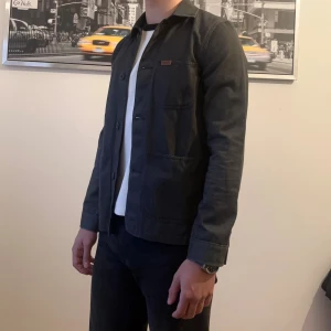 Mörkgrå overshirt från Carhartt - Stilren mörkgrå overshirt från Carhartt med klassisk krage, tre stora utanpåfickor och knäppning framtill. Jackan är i bomull och har en enkel, rak passform som funkar till många olika looks. Perfekt lager-på-lager-plagg för dig som gillar minimalistisk stil.