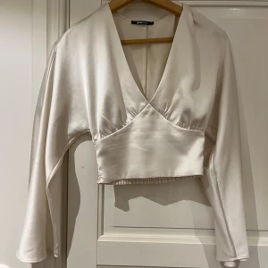 Topp Gina Tricot - Säljer en vit topp från Gina Tricot i glansigt satinliknande material. Toppen är croppad med lång ärm, v-ringning och smockad rygg. Perfekt för dig som gillar trendiga och stilrena plagg med en lyxig känsla. Glansig i färgen!