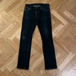 Mörkblå jeans från Nudie Jeans - Snygga mörkblå jeans från Nudie Jeans med klassisk femficksdesign och kontrastsömmar i orange. Bakfickorna har Nudie Jeans signaturbroderi och läderpatch med logga. Jeansen har rak passform och är tillverkade i slitstarkt denimtyg. Ny pris 1600kr